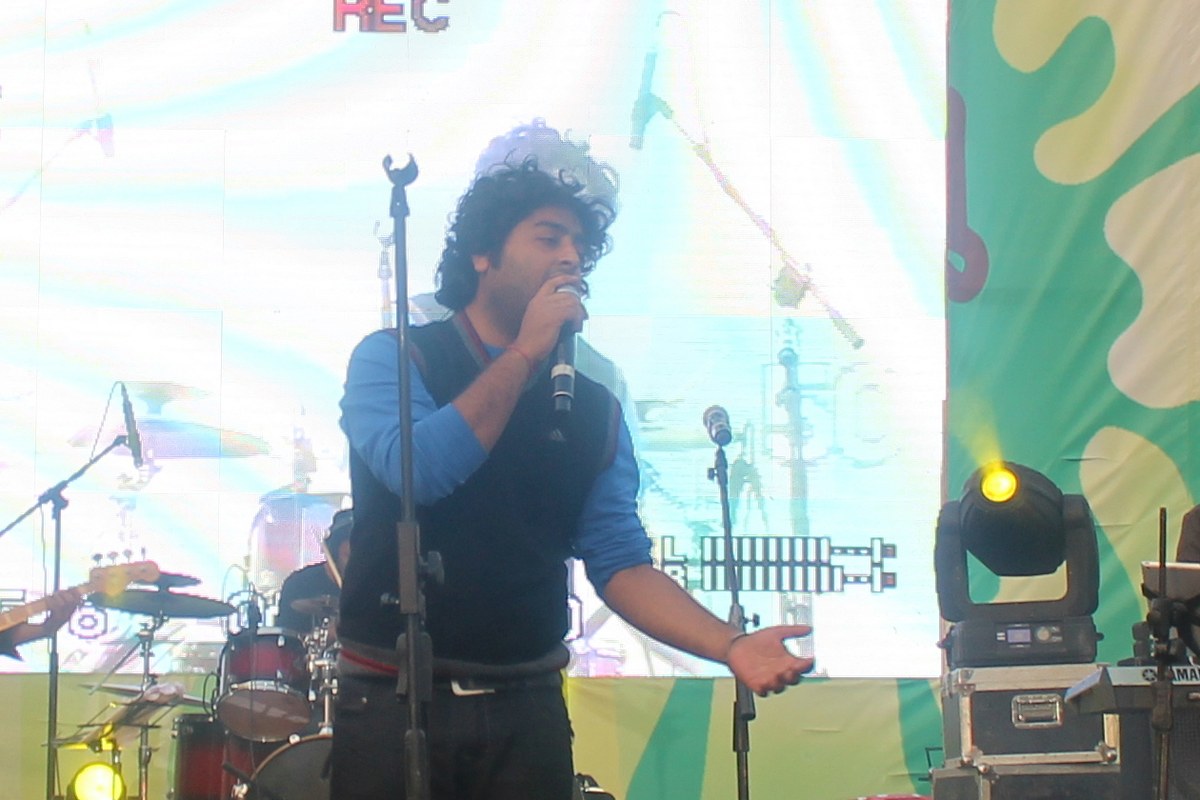 Arijit Singh in a blue shirt and dark waistcoat singing into a microphone on a festival stage.