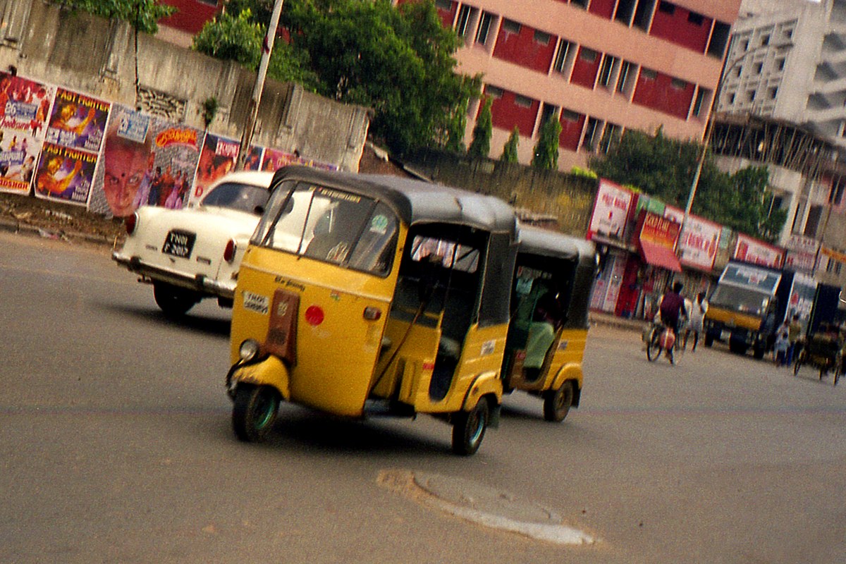 Yellow-and-black autorickshaws driving along a street in Chennai, with a white Ambassador car and shopfronts behind them.