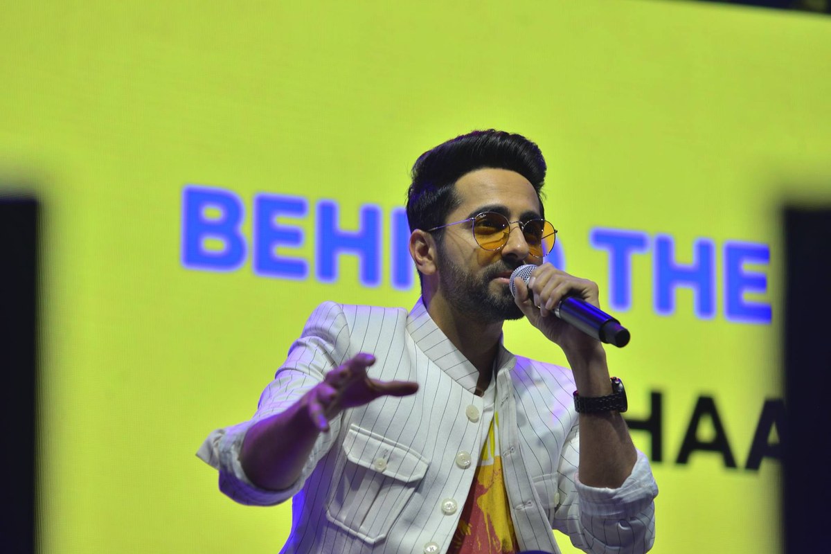 Ayushmann Khurrana, in a white pinstriped jacket and tinted round glasses, speaks into a microphone on a stage with a bright yellow screen behind him.