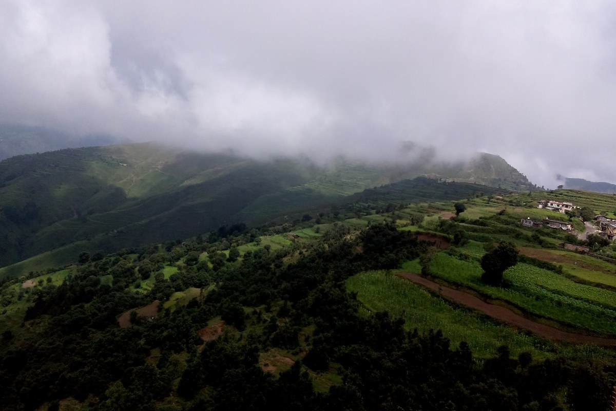 Mist hangs over green terraced hillsides and forest near Chakrata, with a few houses and a dirt track on a ridge to the right.