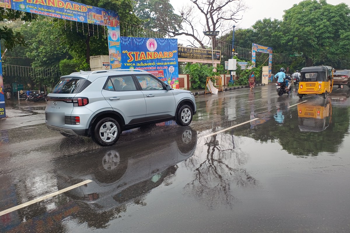 Cars, an auto-rickshaw and motorcyclists on a rain-soaked Anna Salai in Chennai, with puddles and reflections on the road under an overcast sky.