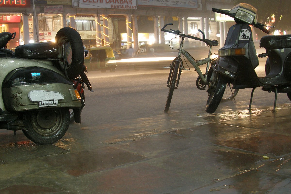 Scooters and a bicycle parked on a wet pavement in heavy rain at night in Connaught Place, New Delhi.