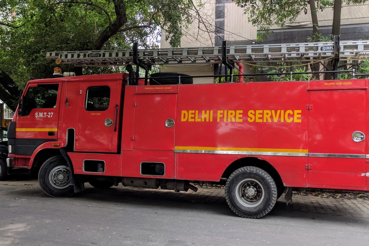 A red Delhi Fire Service fire truck with a ladder on its roof parked on a paved road under trees.