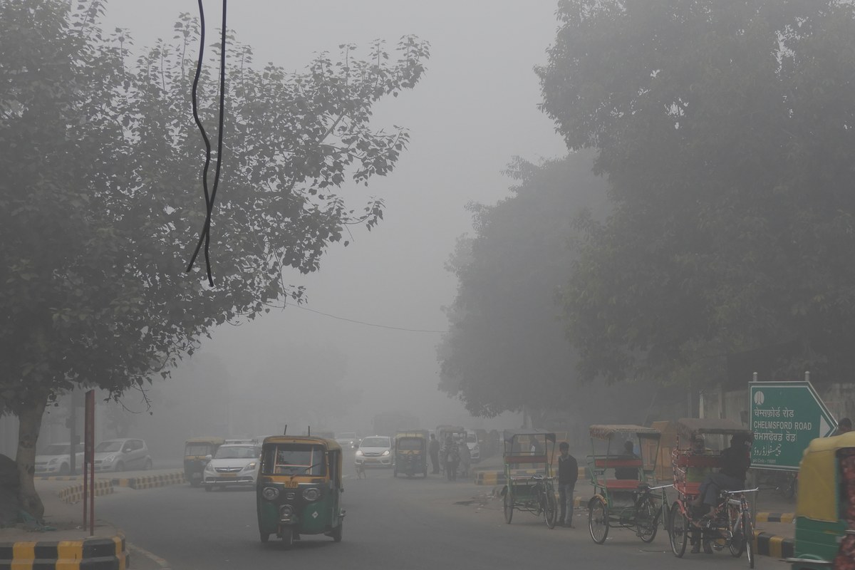 Auto-rickshaws, cars and cycle-rickshaws on a tree-lined New Delhi road shrouded in thick grey smog, with a Chelmsford Road sign on the right.