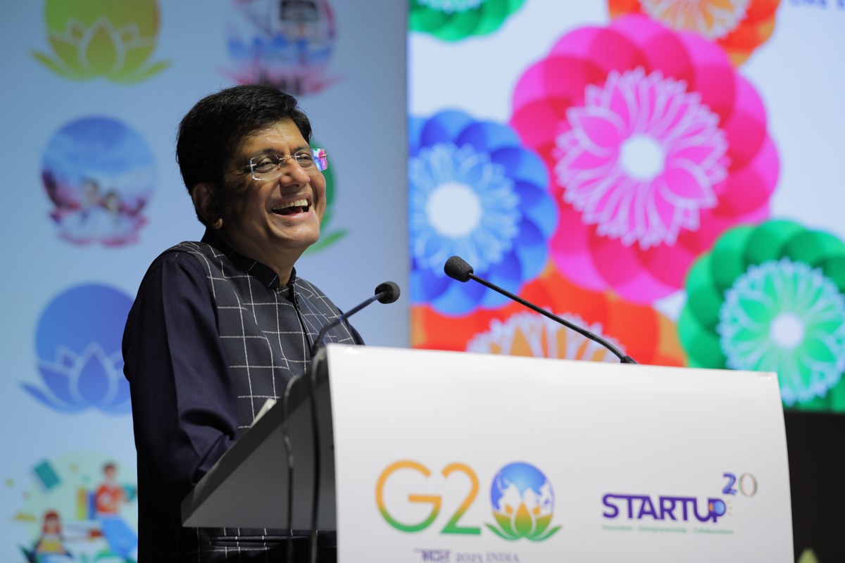 Piyush Goyal, wearing glasses and a dark checked jacket, laughs while speaking at a podium with G20 and Startup20 branding, against a colourful floral backdrop.