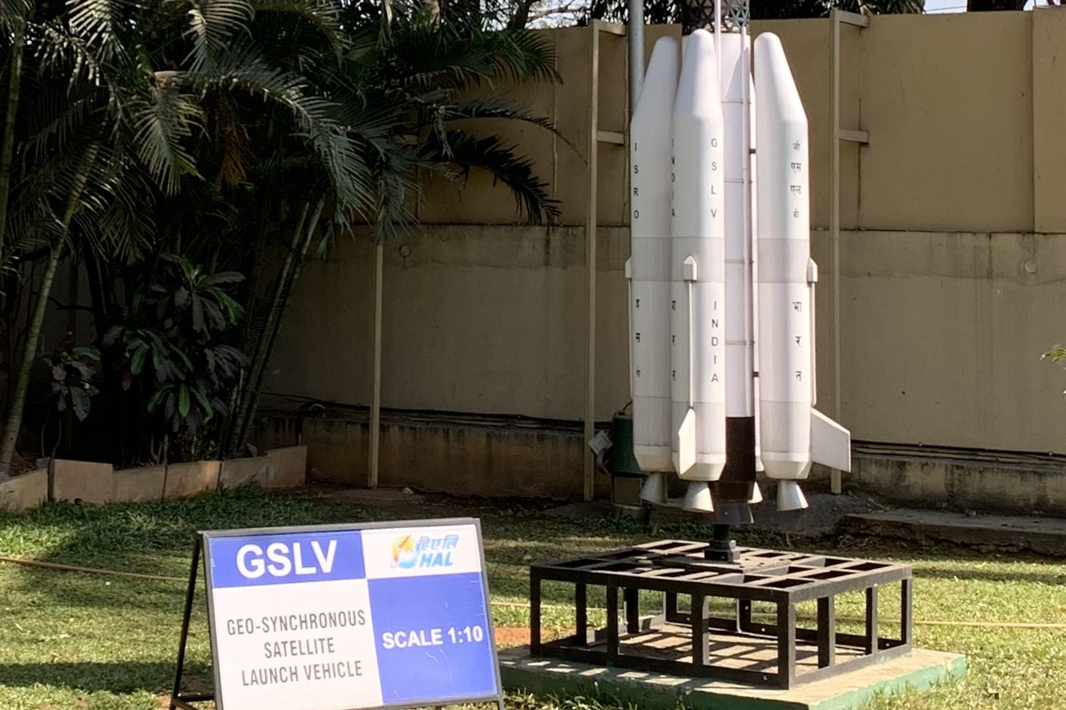 A white scale model of the GSLV rocket on a metal stand on a lawn, next to a blue and white sign reading GSLV, Geo-synchronous Satellite Launch Vehicle, Scale 1:10.