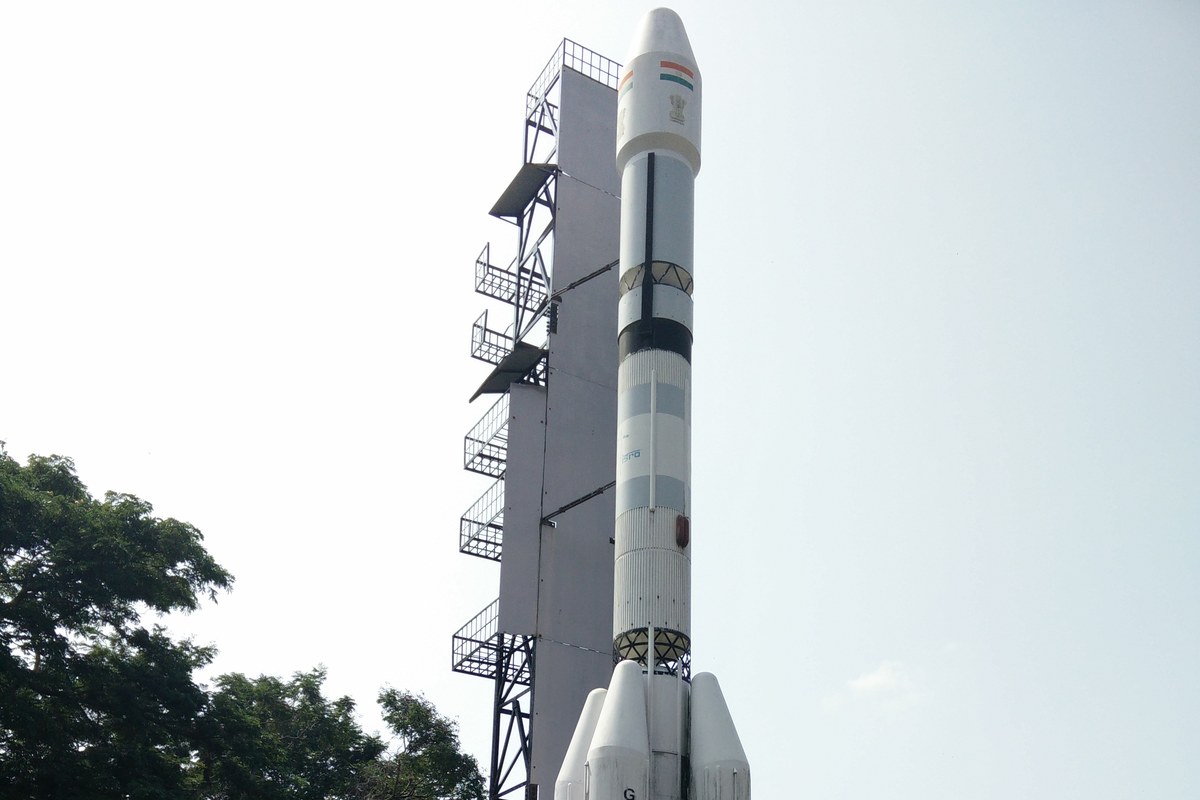 A tall white model of a GSLV rocket with strap-on boosters beside a grey launch tower model, against a pale sky and trees.