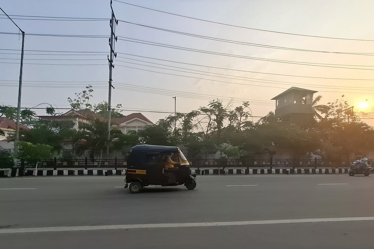 An autorickshaw drives along a wide, quiet road in Guwahati at sunset, with trees, low buildings and power lines along the roadside.
