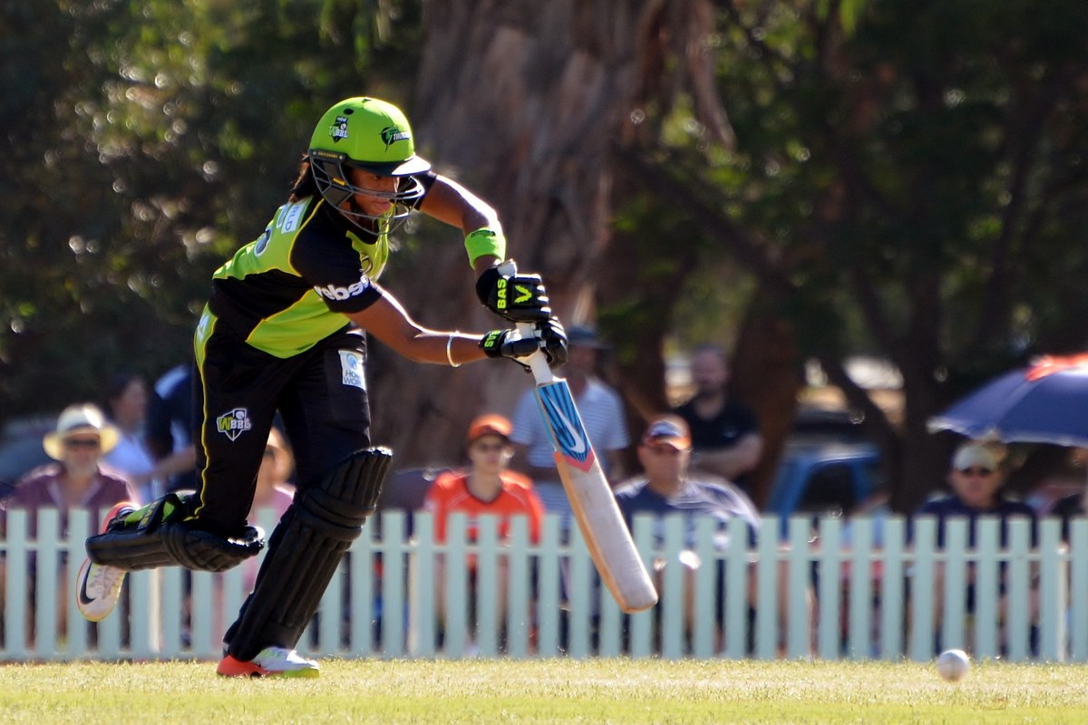 Harmanpreet Kaur in a green Sydney Thunder kit and helmet playing a shot, with spectators behind a white picket fence.