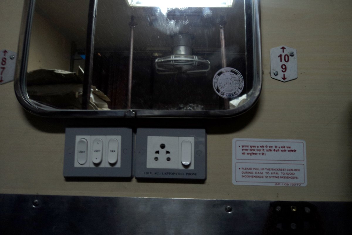 A light switch panel and a socket labelled for laptops and cell phones below a window inside an Indian Railways AC coach.