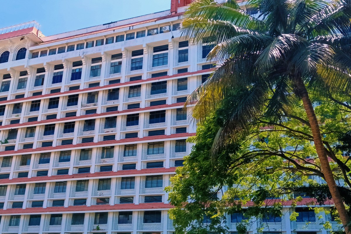 The tall white-and-red High Court of Kerala building in Ernakulam behind a coconut palm and green trees under a blue sky.