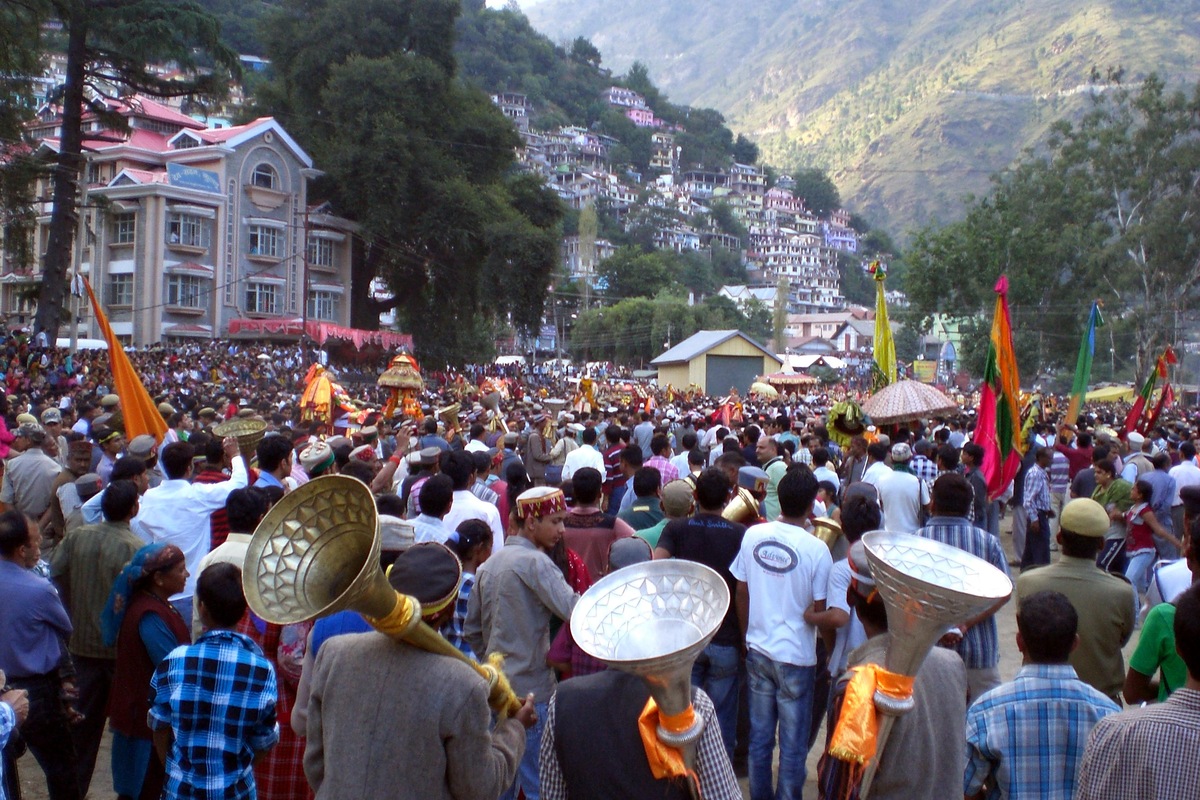 Musicians carrying large brass instruments among a crowded Kullu Dussehra procession, with decorated palanquins and hills beyond.