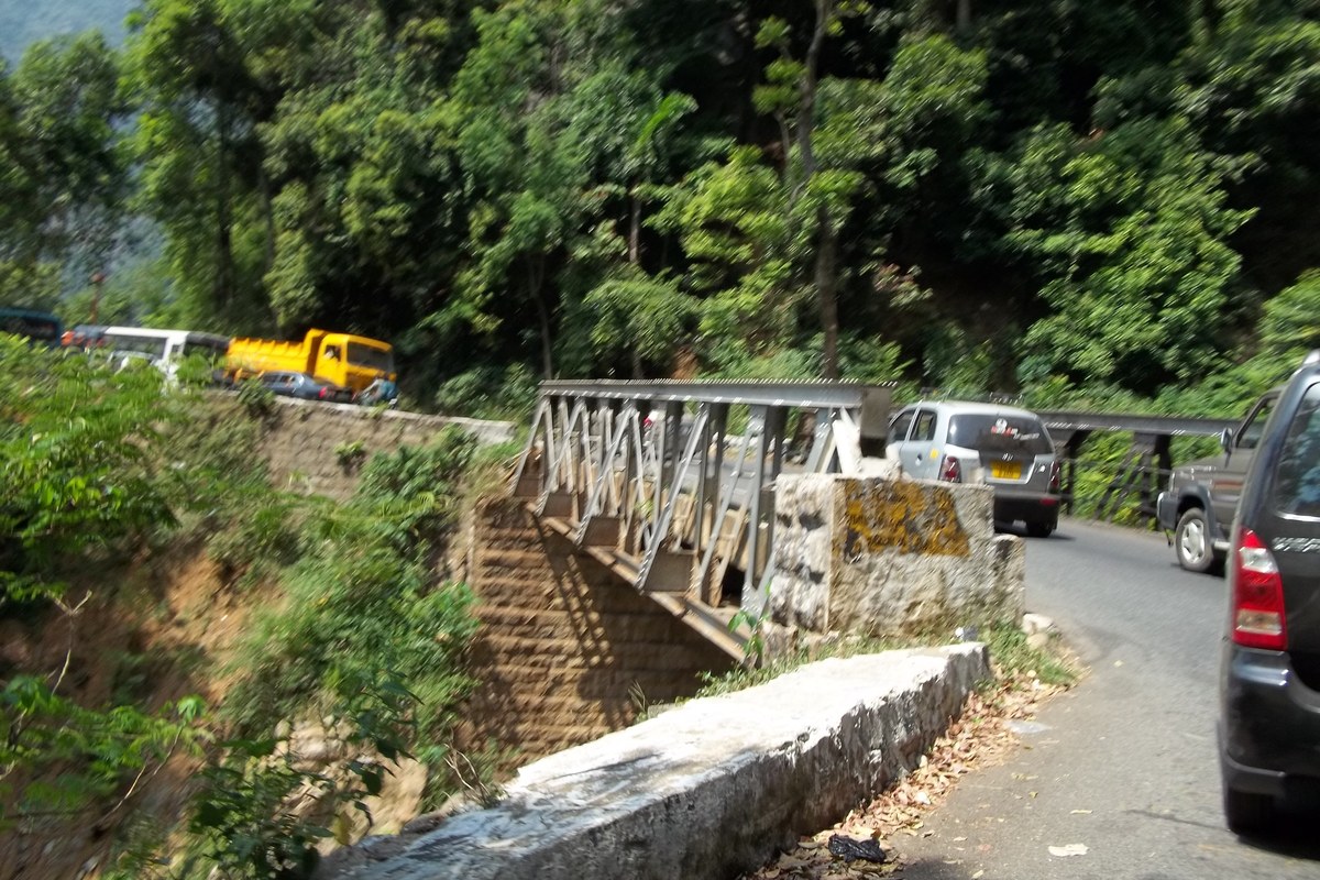 Cars, a bus and a yellow truck on a narrow forested stretch of the Mettupalayam–Coonoor ghat road, beside a small steel bridge and a stone parapet.