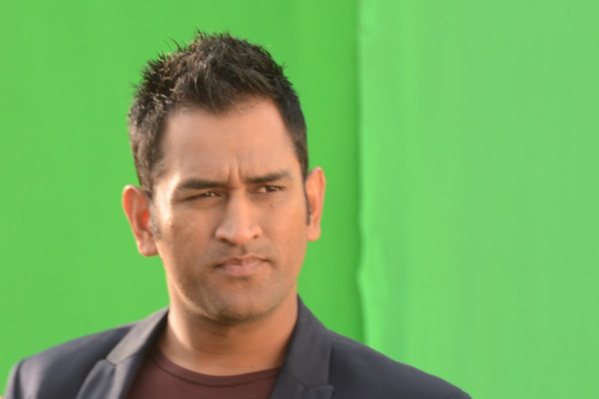 MS Dhoni in a dark blazer looking to his right in front of a green screen.