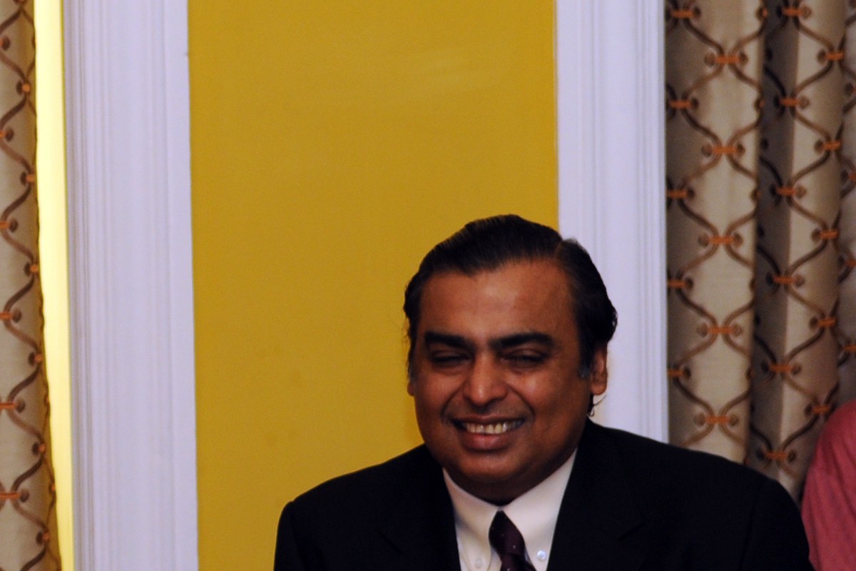 Mukesh Ambani in a dark suit and tie, smiling, seated at a table in front of a yellow wall and patterned curtains.