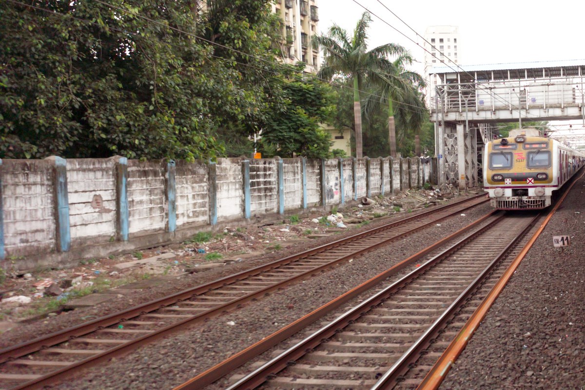 A Mumbai suburban local train approaching on a straight track beside a wall, with trees and a footbridge in the background.