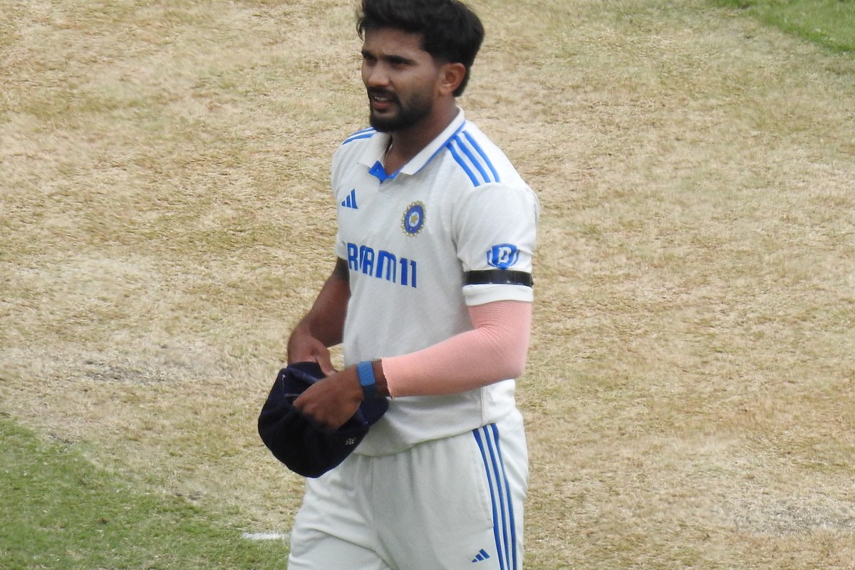 Nitish Kumar Reddy, in India’s white Test kit and a black armband, walks across the outfield holding a navy cap.