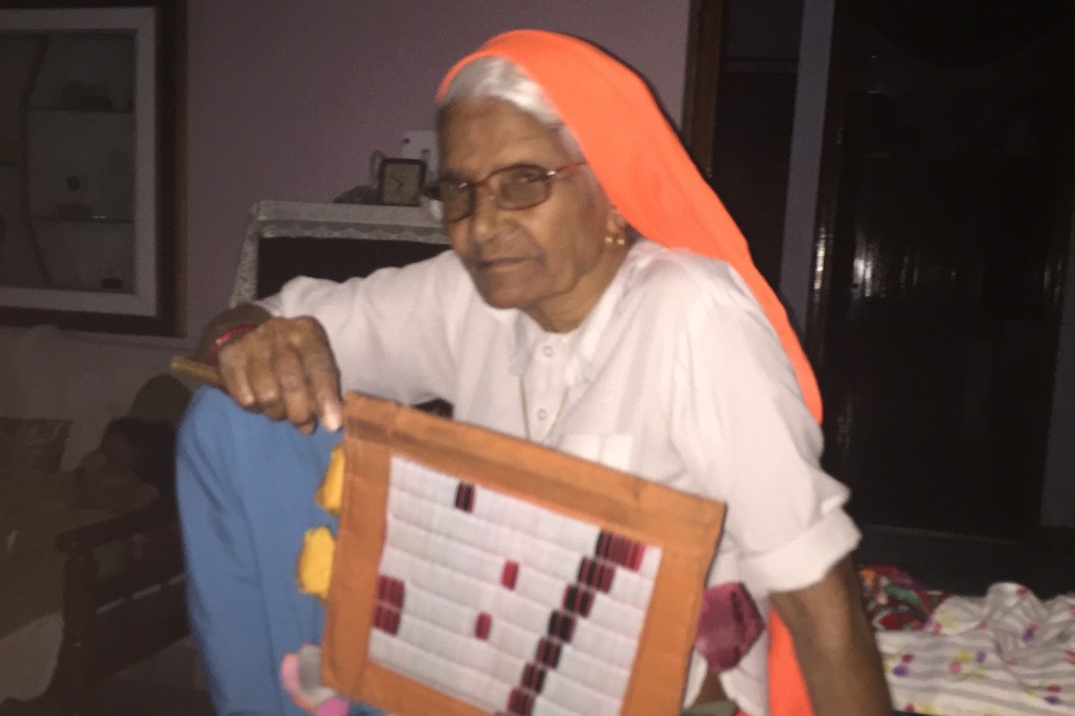 An elderly woman in an orange head covering and glasses sits on a bed holding a traditional hand fan during a power cut in the heat.