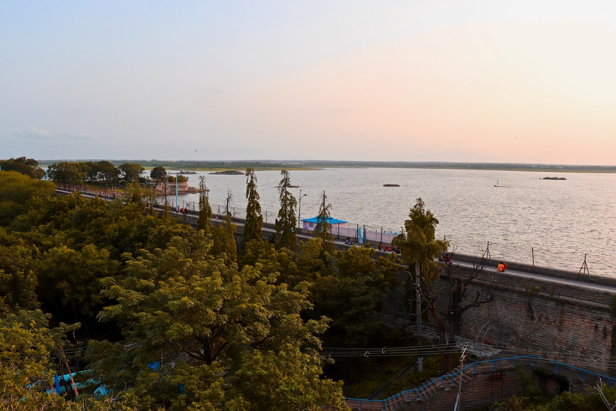 A wide reservoir under a pale evening sky, seen over green trees from the stone retaining wall and road of Osman Sagar near Hyderabad.