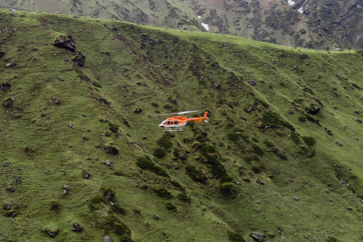 An orange and white helicopter flying low over a green mountain slope with snow-streaked peaks behind.