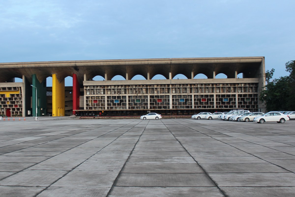The Punjab and Haryana High Court building in Chandigarh, a long concrete structure with a curved roof and tall yellow, green and red pillars, seen across an empty paved plaza.