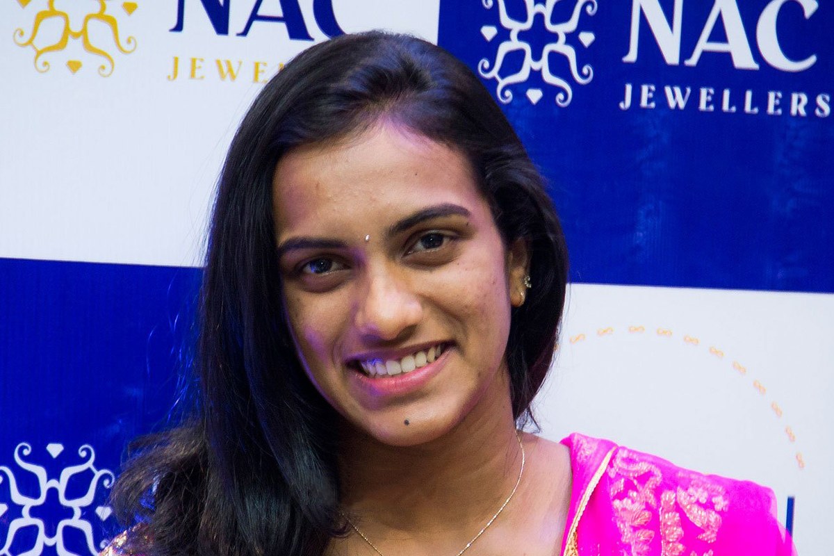 PV Sindhu smiling, wearing a pink outfit with gold embroidery, in front of a blue and white NAC Jewellers backdrop.
