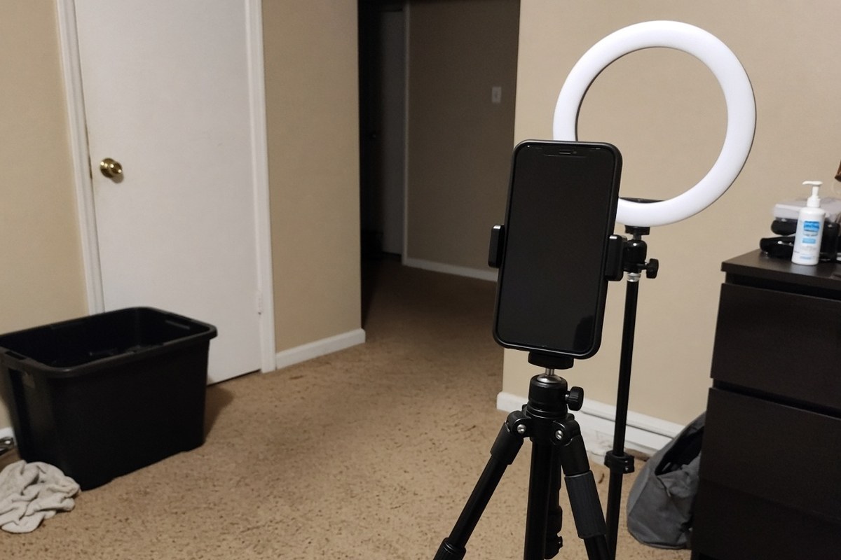 A smartphone on a small tripod beside a switched-on ring light in an empty room, in an AI-generated representative scene of a video-recording set-up.