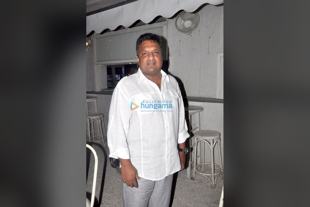 Filmmaker Sanjay Gupta in a white shirt standing in a room with bar stools, shown on a blurred dark background.