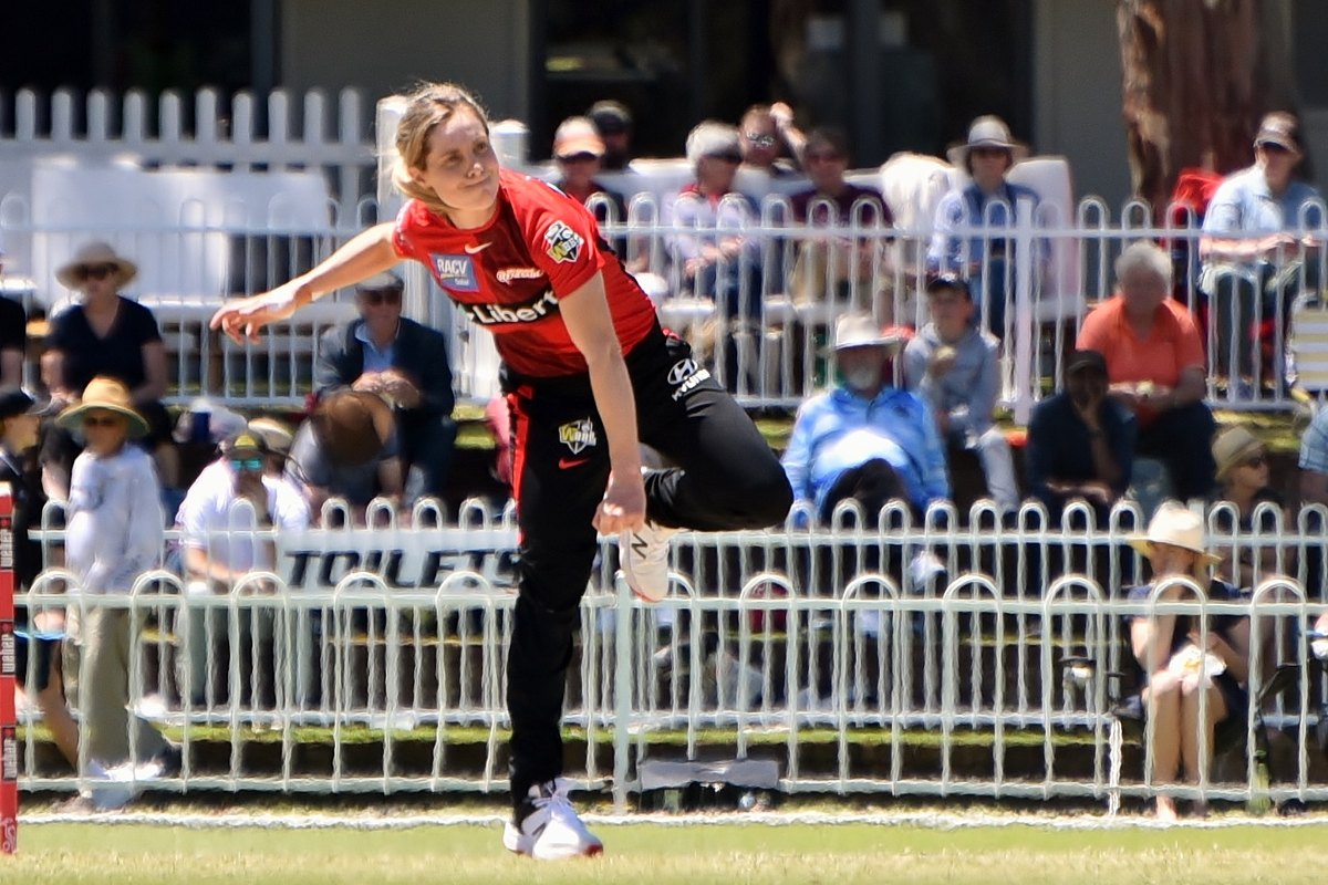 Sophie Molineux in a red Melbourne Renegades shirt follows through after bowling, with spectators behind a white fence.