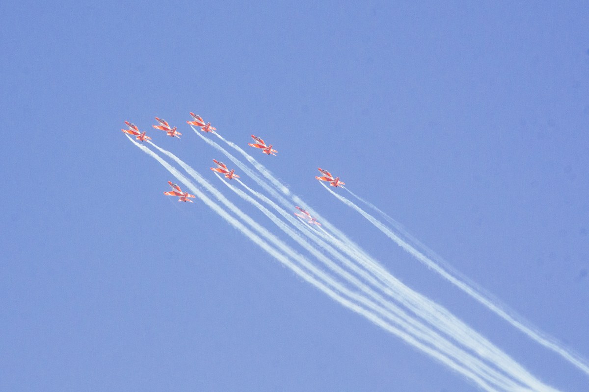 Seven red-and-white Hawk jets of the Surya Kiran aerobatic team flying in formation against a blue sky, trailing white smoke.
