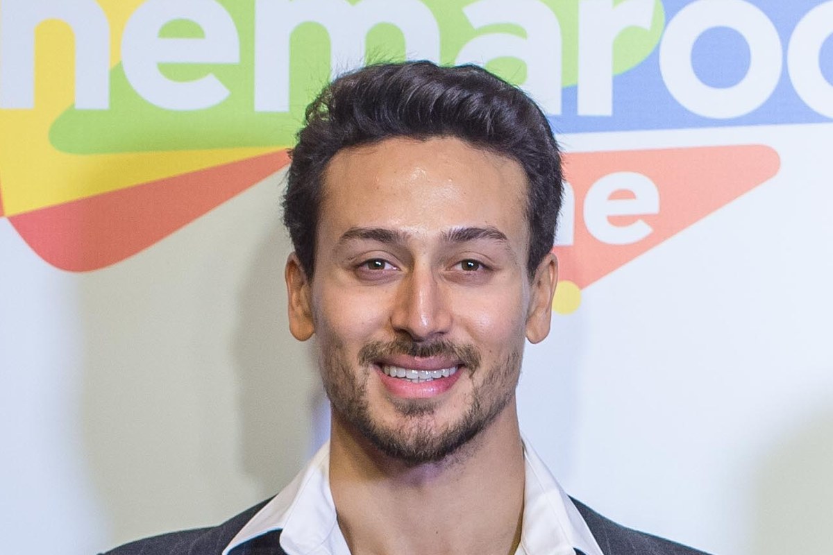 Tiger Shroff smiling in a dark jacket and white shirt in front of a colourful event backdrop.