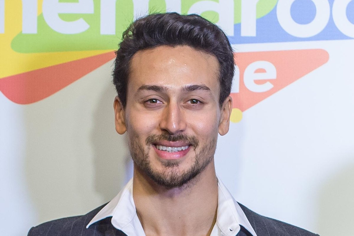 Tiger Shroff smiling, in a dark pinstriped jacket and open-collared white shirt, in front of a colourful event backdrop.