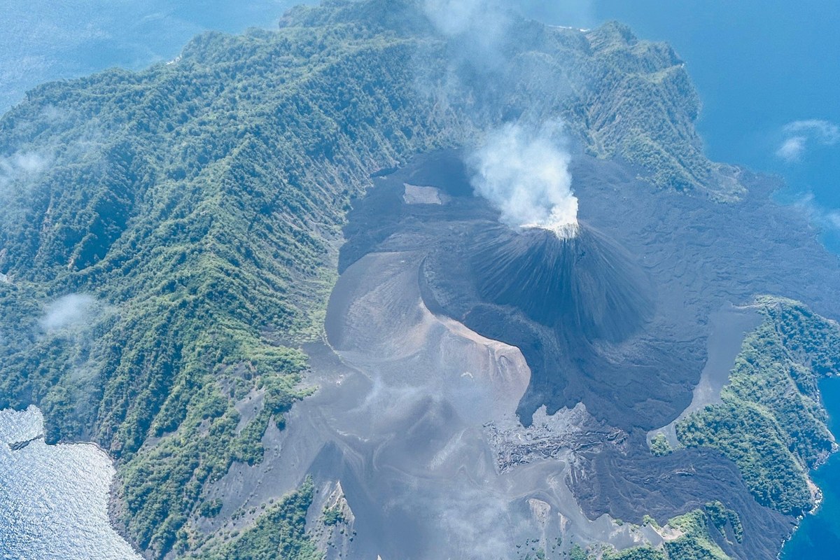 Aerial view of Barren Island, a forested volcanic island with a dark ash cone emitting a white plume of smoke, surrounded by blue sea.