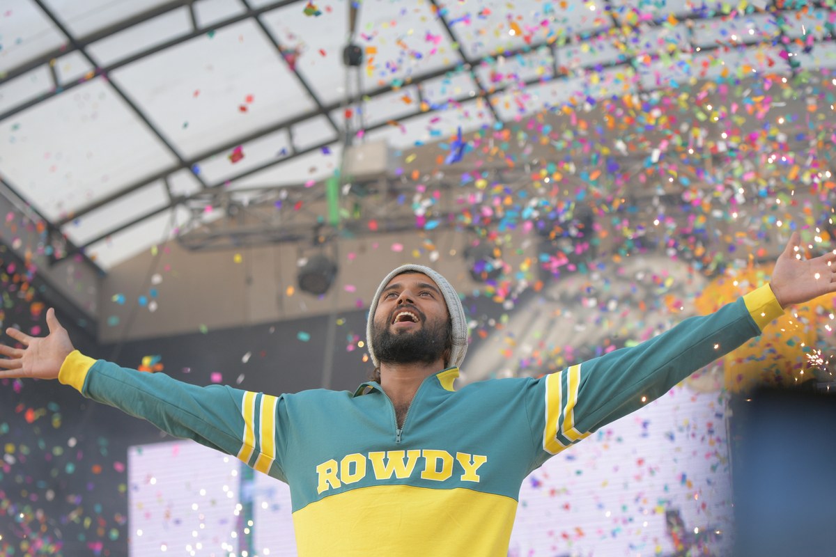 Vijay Deverakonda in a green and yellow ‘Rowdy’ sweatshirt and grey beanie, arms spread wide and smiling as confetti falls around him on stage.